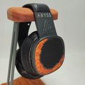 A pair of Abyss over-ear headphones with black leather and orange wood-grain accents, displayed on a wooden stand against a light gray background.