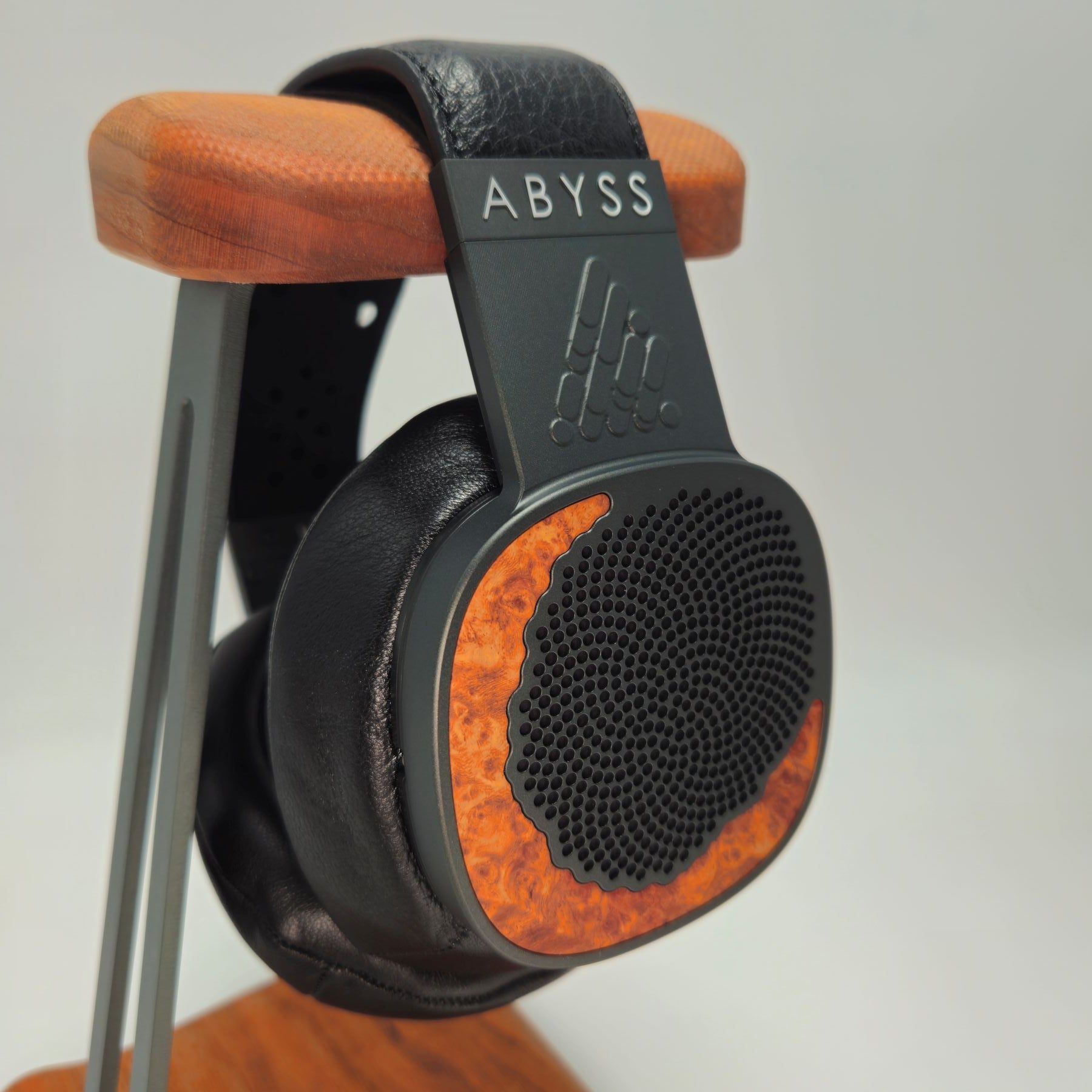 A pair of Abyss over-ear headphones with black leather and orange wood-grain accents, displayed on a wooden stand against a light gray background.