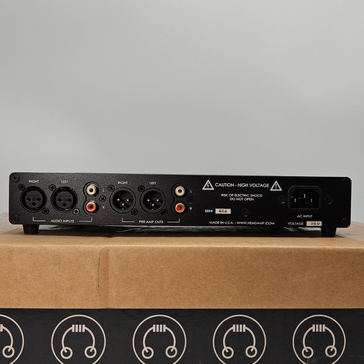 An audio device control panel with various jacks and knobs shown next to its brown cardboard packaging box featuring a black band with the Audiolight logo.