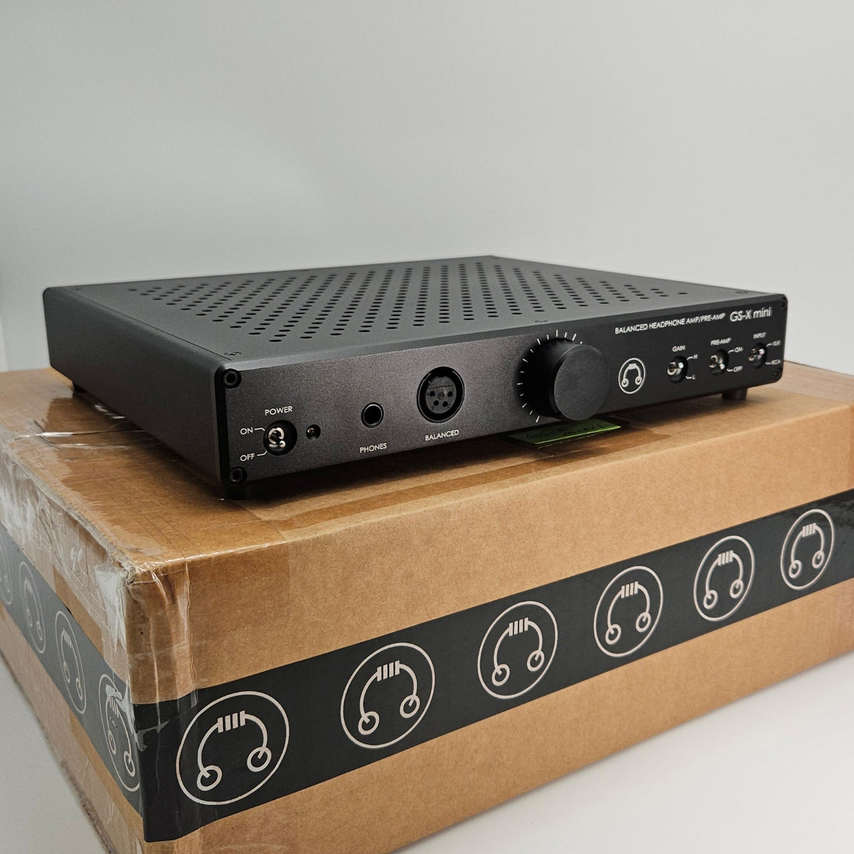 A black audio receiver or amplifier device with a front dial and ports, resting on top of a brown cardboard box with a black headphone-themed band around the middle, photographed against a light gray studio background.